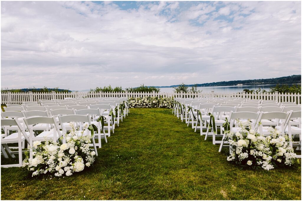 Snohomish Wedding Photographer GSquared Weddings At the picturesque Port Gamble, a stunning summer wedding unfolds. An outdoor ceremony features rows of white chairs facing a floral altar arrangement, with abundant white flowers lining the aisle. The scene overlooks a tranquil body of water under a partly cloudy sky, scattered with greenery. Serving Seattle, Snohomish and Orlando