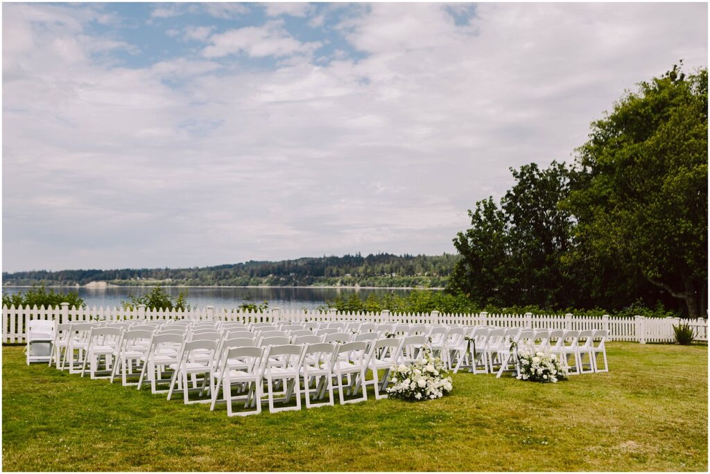 Snohomish Wedding Photographer GSquared Weddings A serene summer wedding setup in Port Gamble features rows of white chairs arranged on a lush green lawn, overlooking a calm lake. White flower arrangements grace the aisles end, while a white picket fence and trees stand proudly under the partly cloudy sky. Serving Seattle, Snohomish and Orlando