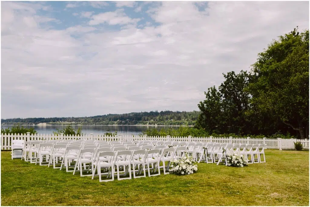 Snohomish Wedding Photographer GSquared Weddings A serene summer wedding setup in Port Gamble features rows of white chairs arranged on a lush green lawn, overlooking a calm lake. White flower arrangements grace the aisles end, while a white picket fence and trees stand proudly under the partly cloudy sky. Serving Seattle, Snohomish and Orlando
