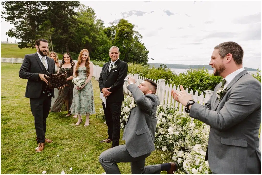 Snohomish Wedding Photographer GSquared Weddings A man in a gray suit kneels, holding a ring box during a beautiful Port Gamble summer wedding. Another man in a suit stands clapping, surrounded by smiling bridesmaids in floral dresses and two other groomsmen. They are outdoors with trees and a white fence in the background. Serving Seattle, Snohomish and Orlando