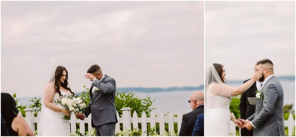 Snohomish Wedding Photographer GSquared Weddings A bride and groom stand outdoors by a white picket fence in Port Gamble. The bride is in a white gown, holding a bouquet, while the groom is in a gray suit. Both are smiling and adjusting their hair, embodying the joy of their summer wedding against a backdrop of cloudy sky and distant water. Serving Seattle, Snohomish and Orlando