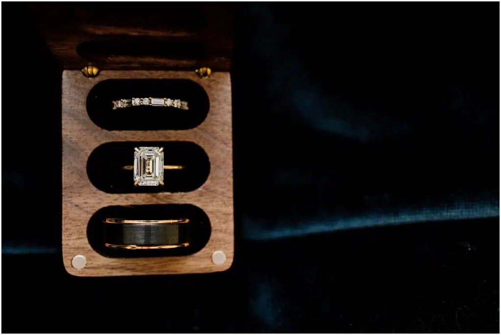 Snohomish Wedding Photographer GSquared Weddings A wooden box reveals three exquisite rings: a thin diamond band on top, a central emerald-cut diamond ring, and a wider band below. Perfect for a Port Gamble summer wedding, the open box rests against a dark, textured background that enhances their sparkle. Serving Seattle, Snohomish and Orlando