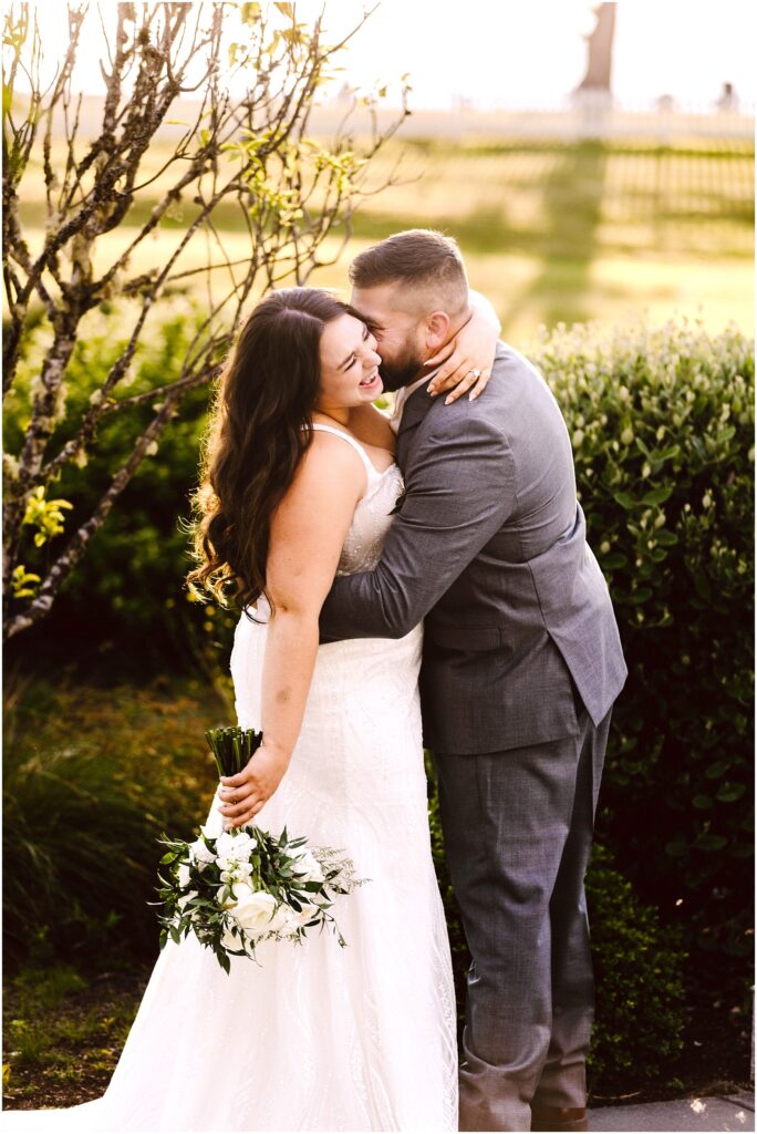 Snohomish Wedding Photographer GSquared Weddings At a picturesque summer wedding in Port Gamble, a bride in a white dress holds a bouquet and smiles while embracing a groom in a gray suit. They stand outdoors near a hedge, with sunlight and lush greenery enhancing the romantic scene. Serving Seattle, Snohomish and Orlando