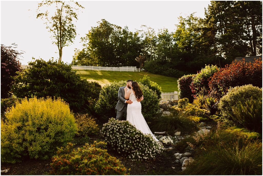 Snohomish Wedding Photographer GSquared Weddings Amidst a lush gardens vibrant greenery and colorful bushes, a bride and groom share a kiss as the sun casts its warm glow over their summer wedding in Port Gamble. Serving Seattle, Snohomish and Orlando