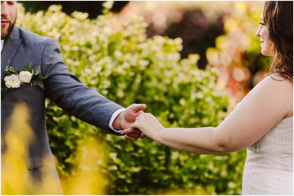 Snohomish Wedding Photographer GSquared Weddings A bride and groom are holding hands outdoors at their summer wedding in Port Gamble. The groom is dressed in a gray suit with a boutonniere, and the bride is wearing a white dress. They are surrounded by lush greenery and blurred colorful flowers in the background. Serving Seattle, Snohomish and Orlando