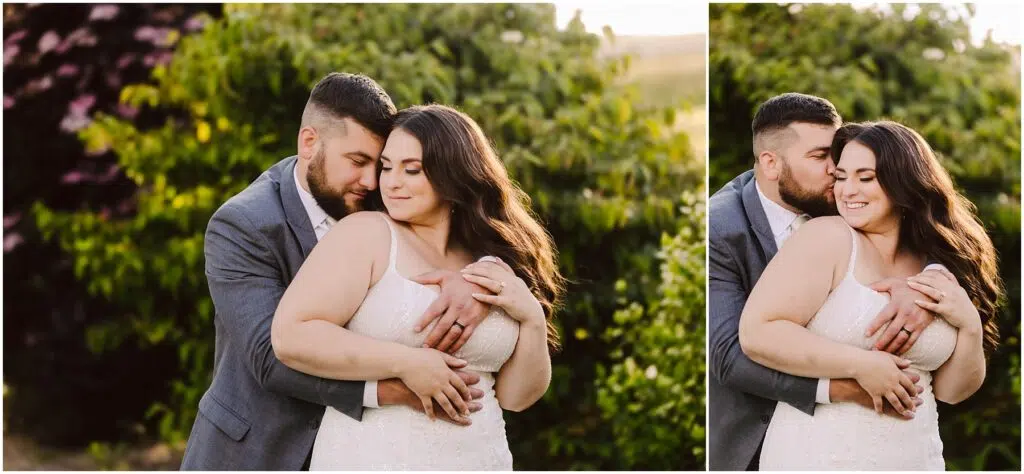 Snohomish Wedding Photographer GSquared Weddings In the heart of Port Gamble, amidst lush greenery, a couple embraces warmly. The man, in a gray suit, hugs the woman in her white dress from behind. Their content expressions and the soft summer wedding sunlight illuminate the serene scene. Serving Seattle, Snohomish and Orlando