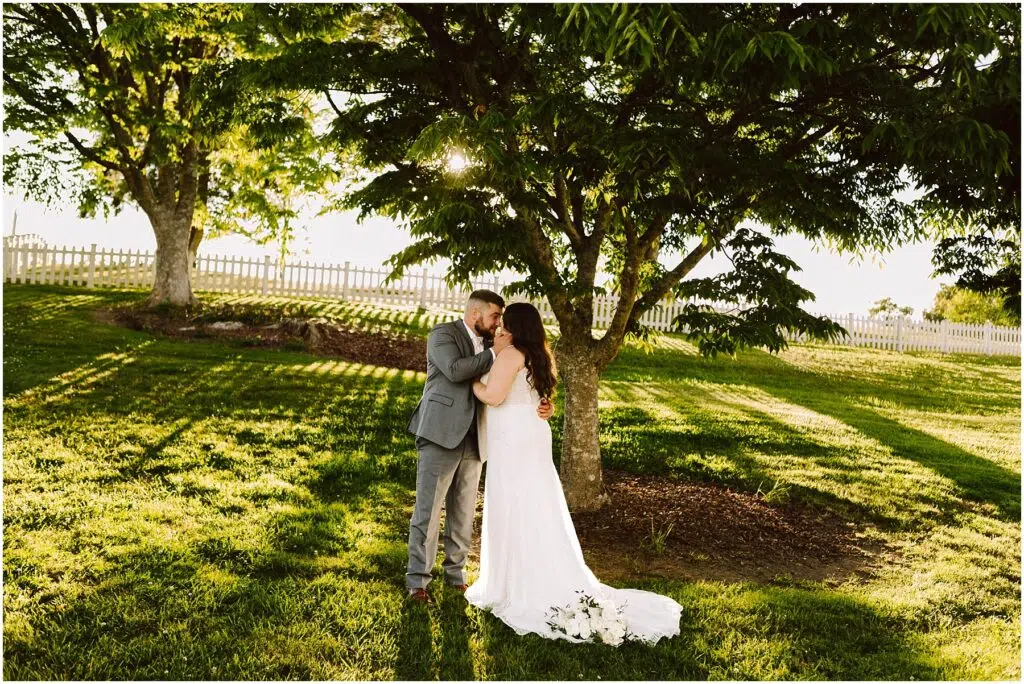 Snohomish Wedding Photographer GSquared Weddings A couple stands under a large tree in a sunlit Port Gamble garden. The woman wears a white wedding dress, and the man is in a gray suit. Embracing lovingly, theyre enveloped by sunlight filtering through the leaves, embodying the essence of a perfect summer wedding day. Serving Seattle, Snohomish and Orlando