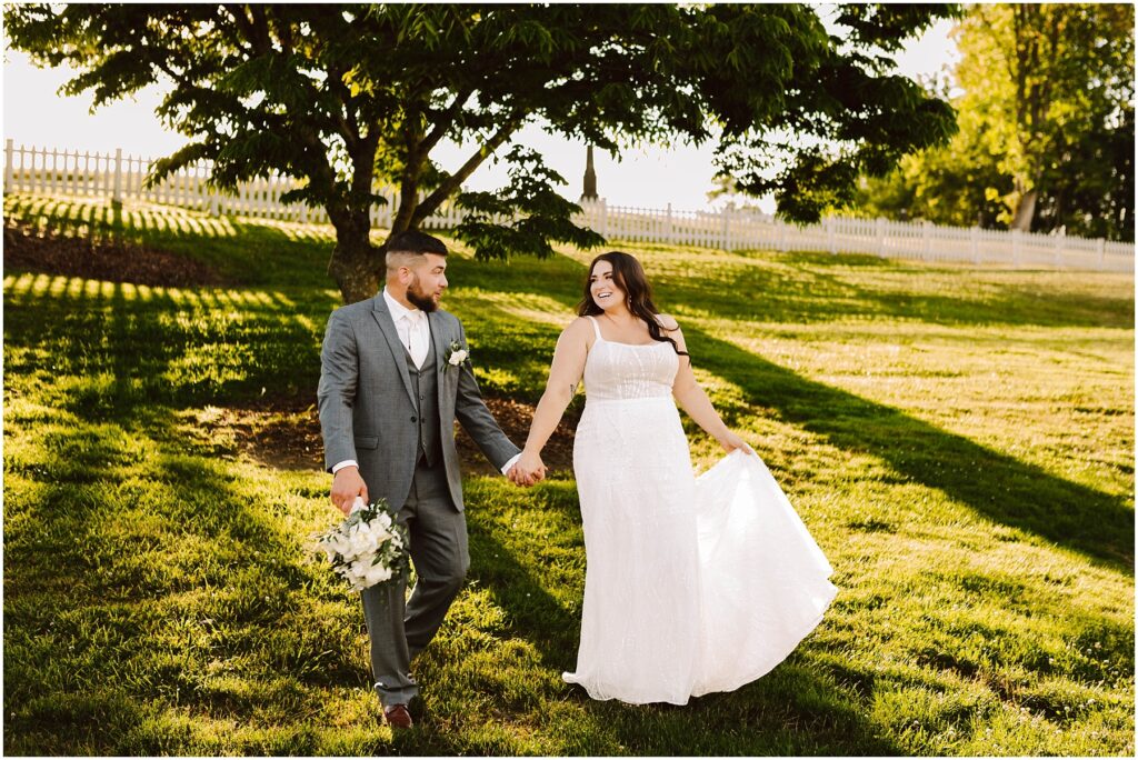 Snohomish Wedding Photographer GSquared Weddings A smiling bride and groom hold hands, strolling through a grassy field at their Port Gamble summer wedding. The groom, in a gray suit with a bouquet, and the bride, lifting her white dresss hem, are framed by sunlit trees and a charming white fence in the background. Serving Seattle, Snohomish and Orlando