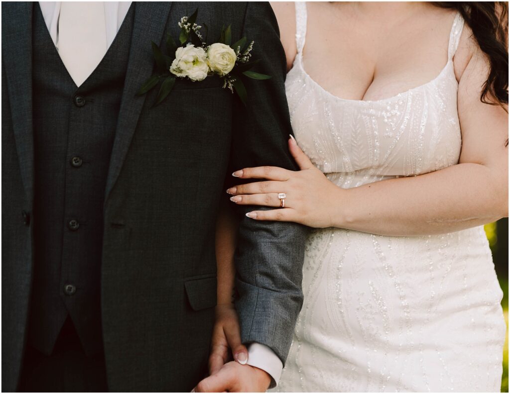 Snohomish Wedding Photographer GSquared Weddings In a close-up of their Port Gamble summer wedding, the newlywed couple stands arm in arm. The groom dons a dark suit with a boutonniere, and the brides white, beaded dress highlights her wedding ring as they hold hands, greenery framing the scene. Serving Seattle, Snohomish and Orlando