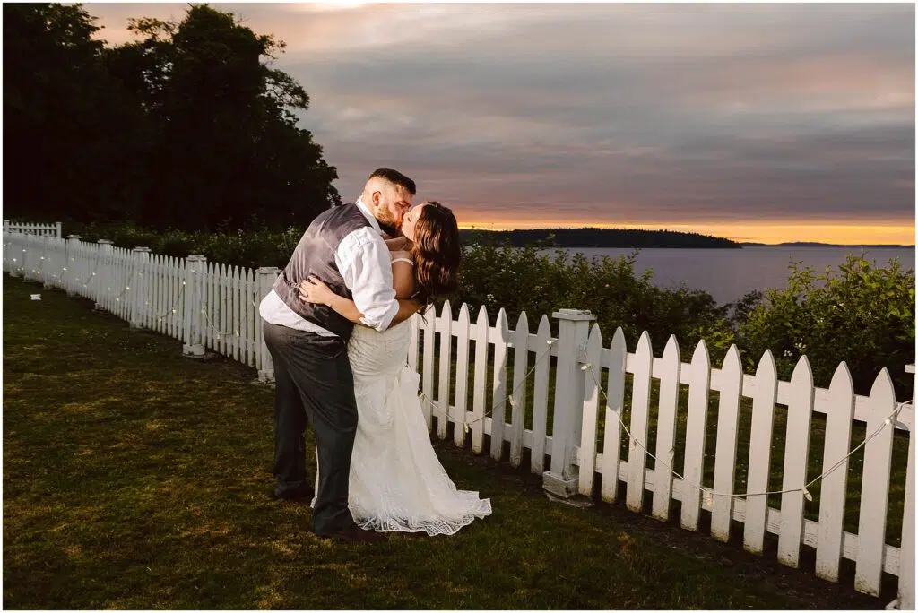 Snohomish Wedding Photographer GSquared Weddings A couple in wedding attire embraces and kisses beside a white picket fence at their summer wedding in Port Gamble, with a picturesque lake and sunset in the background. The evening sky displays shades of purple and orange. Serving Seattle, Snohomish and Orlando