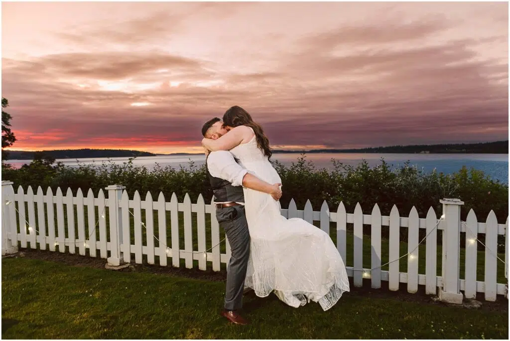 Snohomish Wedding Photographer GSquared Weddings A couple embraces joyfully in front of a white picket fence during sunset at their Summer Wedding. The sky, painted with hues of pink and orange over a calm lake in Port Gamble, adds a romantic backdrop to their celebration. The man lifts the woman, who wears a white dress. Serving Seattle, Snohomish and Orlando