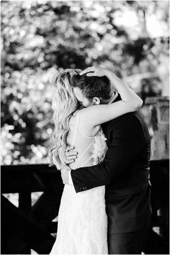 Snohomish Wedding Photographer GSquared Weddings A bride and groom embrace in a heartfelt hug amidst the scenic beauty of a winery. The bride, in an intricate lace gown with long hair cascading down her back, and the groom, in a dark suit, are framed by soft, blurred greenery. Black and white photo. Serving Seattle, Snohomish and Orlando