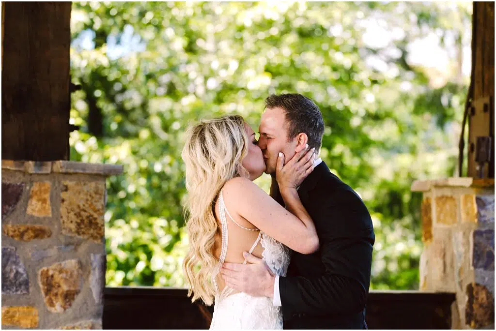 Snohomish Wedding Photographer GSquared Weddings A couple dressed in wedding attire shares a kiss under a stone and wood pavilion at a picturesque winery. The bride, with long blonde hair, and the groom in a black suit, are bathed in sunlight filtering through lush foliage, creating an enchanting and romantic setting. Serving Seattle, Snohomish and Orlando