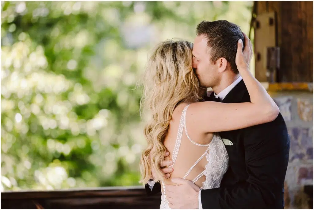Snohomish Wedding Photographer GSquared Weddings A couple in wedding attire shares a kiss outdoors at a charming winery. The groom, in a black suit, embraces the bride, who wears a white gown with lace details. Lush green foliage intermingles with the vineyard backdrop as the brides hand rests on the grooms shoulder. Serving Seattle, Snohomish and Orlando