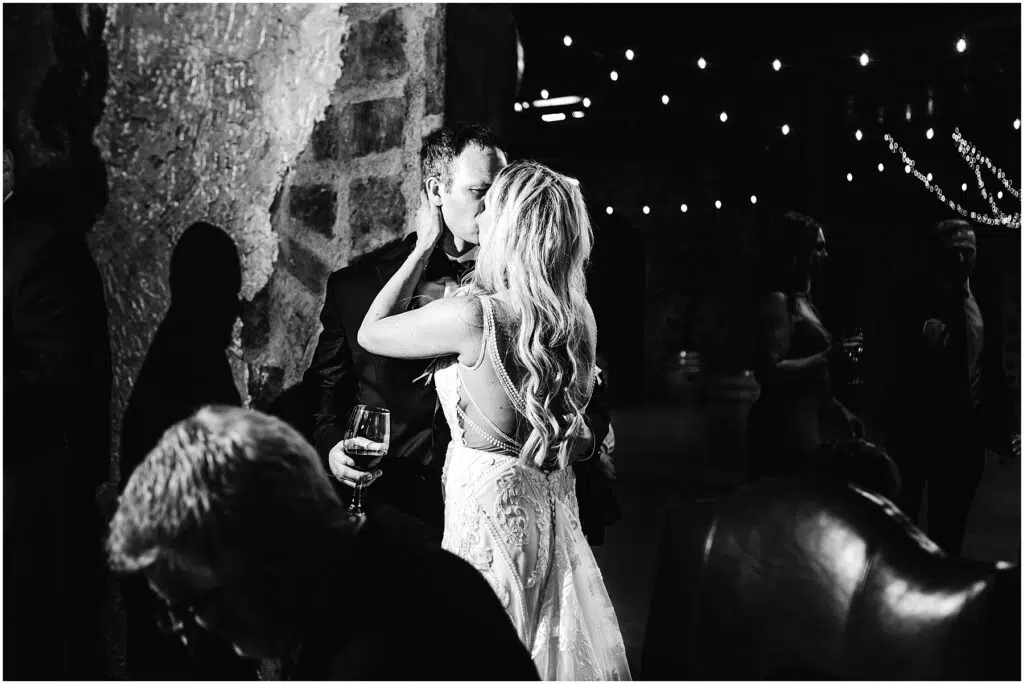 Snohomish Wedding Photographer GSquared Weddings A couple kisses tenderly at a dimly lit winery event. The woman, in a detailed gown, holds a wine glass and faces a stone wall. String lights create a cozy atmosphere, with others talking in the background. Black and white photo. Serving Seattle, Snohomish and Orlando