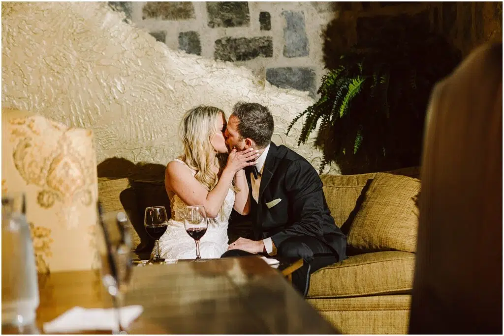 Snohomish Wedding Photographer GSquared Weddings A couple in formal attire kisses while seated on a couch, surrounded by the cozy ambiance of a rustic winery. A stone wall and lush plant provide the backdrop, as two glasses of red wine sit invitingly on the table before them, enhancing the intimate moment. Serving Seattle, Snohomish and Orlando