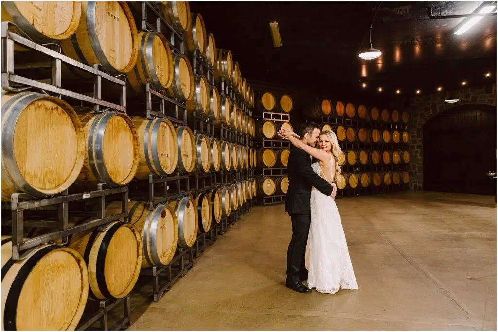 Snohomish Wedding Photographer GSquared Weddings A couple embraces while dancing in the winerys cellar, surrounded by stacks of wooden barrels. The woman is wearing a white dress, and the man is in a suit. Warm lighting creates a cozy atmosphere that perfectly complements the vineyards enchanting charm. Serving Seattle, Snohomish and Orlando