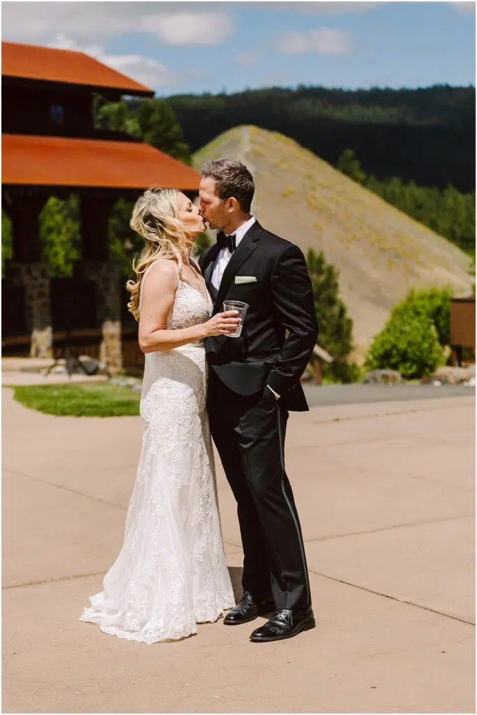 Snohomish Wedding Photographer GSquared Weddings A bride in a white lace dress and a groom in a black tuxedo kiss outdoors at a picturesque winery. The bride holds a drink as they stand on a paved area, with charming buildings and lush, hilly vineyard landscapes under the partly cloudy sky. Serving Seattle, Snohomish and Orlando