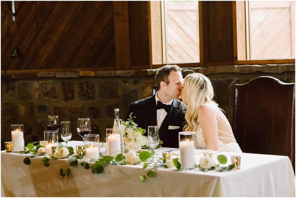 Snohomish Wedding Photographer GSquared Weddings A couple in formal attire kisses at a wedding reception table adorned with white flowers, candles, and greenery. They are seated in front of wooden walls and large windows of a charming winery, with wine glasses and a bottle gracing the table. Serving Seattle, Snohomish and Orlando