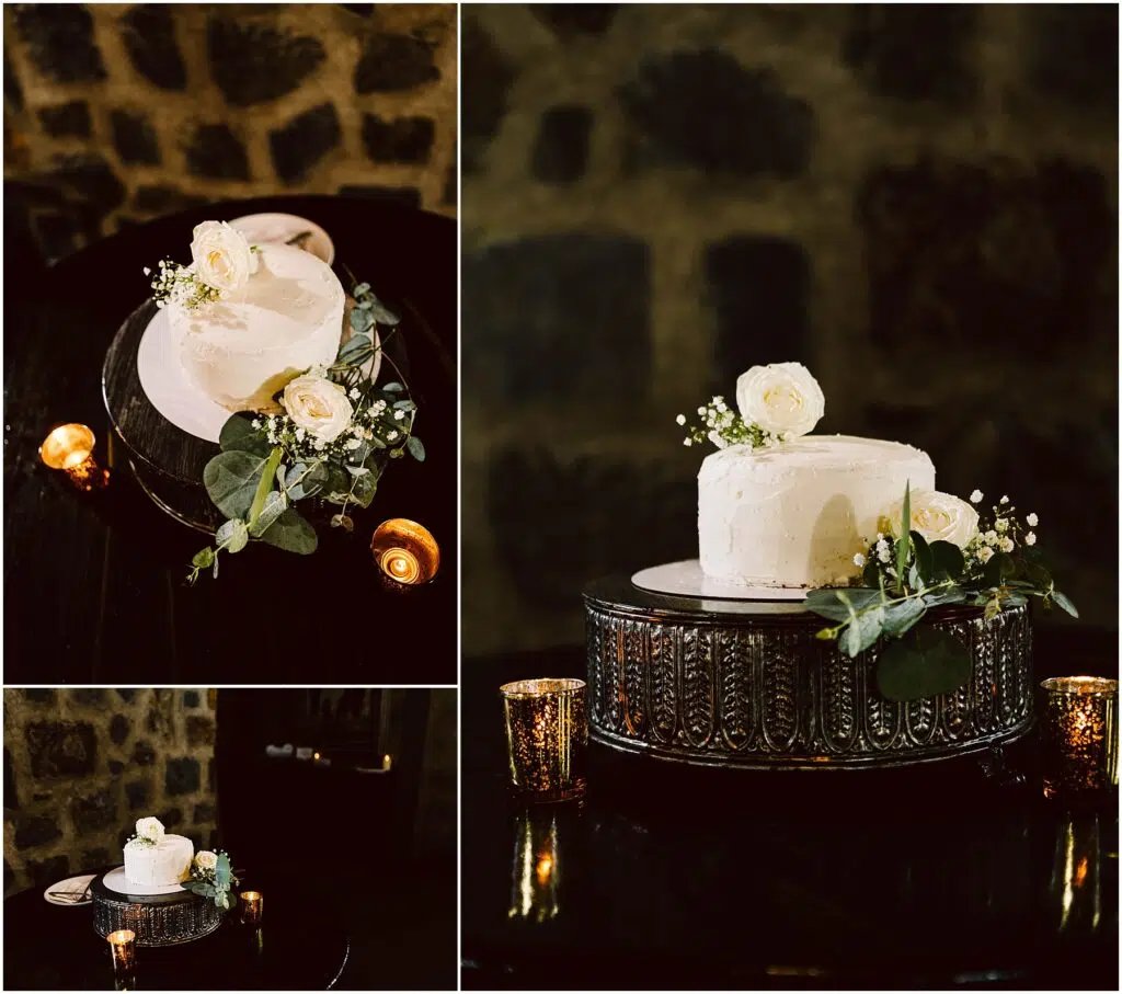 Snohomish Wedding Photographer GSquared Weddings A two-tiered white frosted cake adorned with white roses and greenery sits elegantly on an ornate stand, reminiscent of a winerys rustic charm. The cake is surrounded by small candles, set against a dark stone wall background. There are close-up and angled views of the cake. Serving Seattle, Snohomish and Orlando