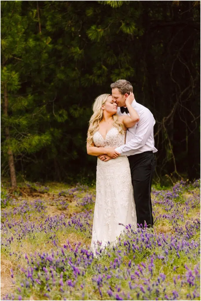 Snohomish Wedding Photographer GSquared Weddings A couple embraces in a field of purple flowers near a serene winery. The man, dressed in a white shirt and black pants, kisses the forehead of the woman in a white lace dress. They both have blonde hair and look content surrounded by trees and grapevines. Serving Seattle, Snohomish and Orlando
