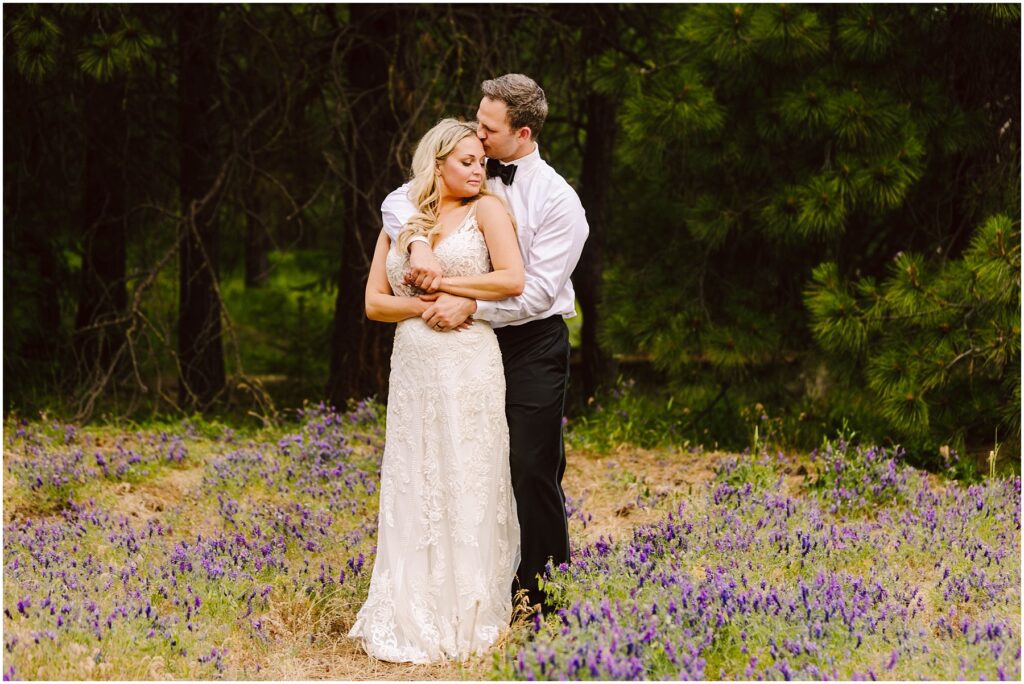 Snohomish Wedding Photographer GSquared Weddings A couple stands in a field of purple flowers near a quaint winery, surrounded by trees. The man, in a white shirt and black pants, embraces the woman, who wears a white lace dress. They both appear serene and content. Serving Seattle, Snohomish and Orlando