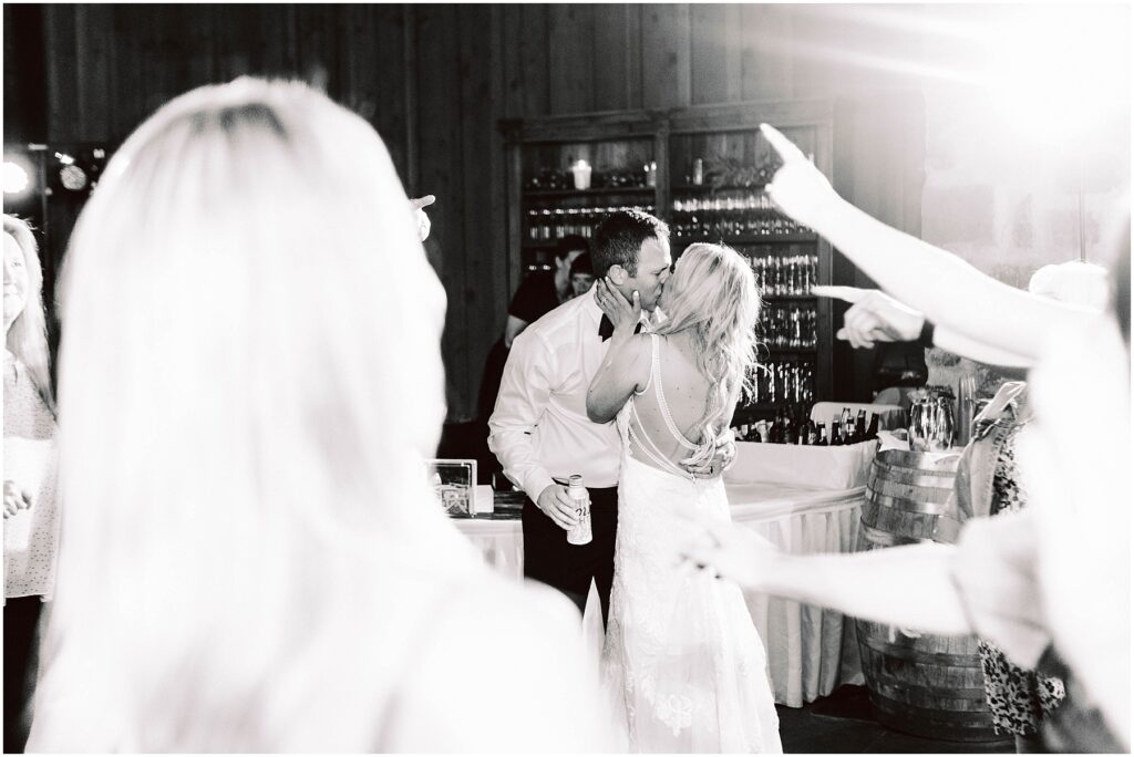 Snohomish Wedding Photographer GSquared Weddings A couple shares a kiss on the dance floor at a wedding reception in a rustic winery. The groom holds a drink, while guests around them celebrate with raised hands. The brides dress features a detailed back design, adding elegance to the festive atmosphere. Serving Seattle, Snohomish and Orlando