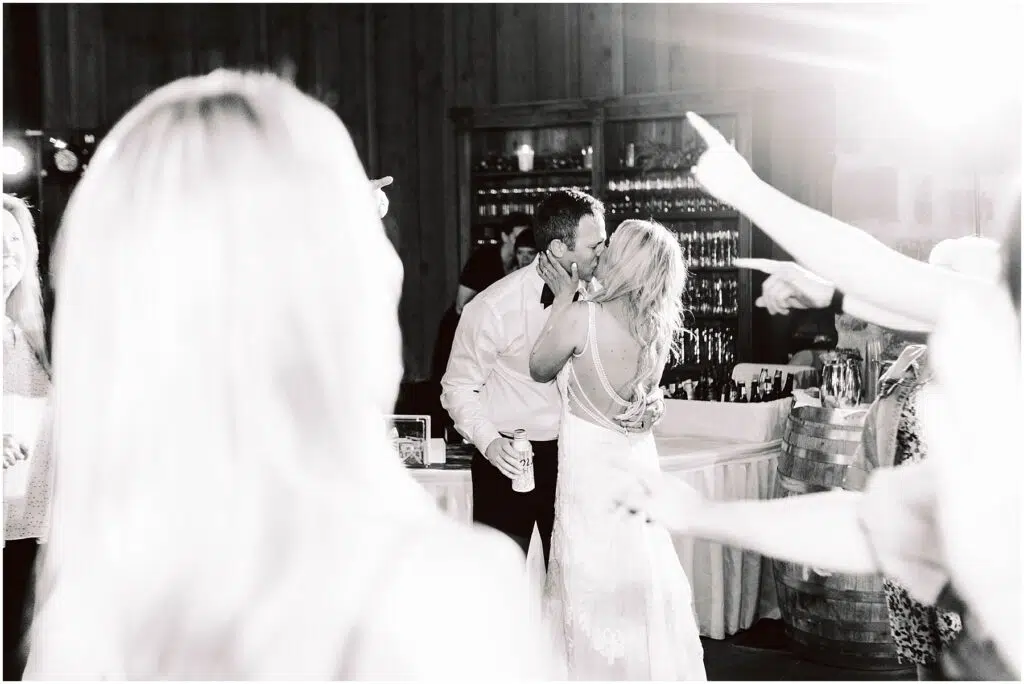 Snohomish Wedding Photographer GSquared Weddings A couple shares a kiss on the dance floor at a wedding reception in a rustic winery. The groom holds a drink, while guests around them celebrate with raised hands. The brides dress features a detailed back design, adding elegance to the festive atmosphere. Serving Seattle, Snohomish and Orlando