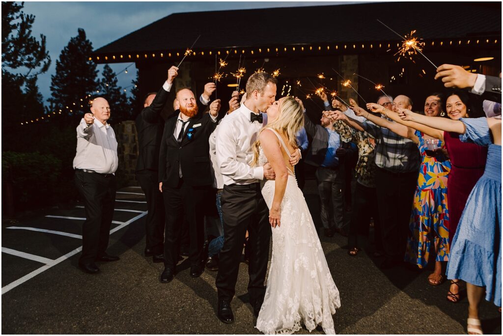 Snohomish Wedding Photographer GSquared Weddings A newlywed couple shares a kiss amid cheering guests holding sparklers at a charming winery. The bride dazzles in a white lace gown, the groom in a black suit. The scene is festive, set against the soft glow of twinkling string lights and lush vineyard rows. Serving Seattle, Snohomish and Orlando
