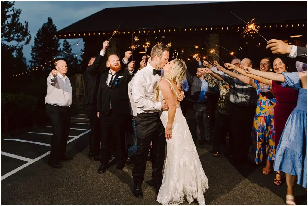 Snohomish Wedding Photographer GSquared Weddings A newlywed couple shares a kiss amid cheering guests holding sparklers at a charming winery. The bride dazzles in a white lace gown, the groom in a black suit. The scene is festive, set against the soft glow of twinkling string lights and lush vineyard rows. Serving Seattle, Snohomish and Orlando