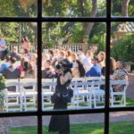Snohomish Wedding Photographer GSquared Weddings A wedding photographer stands on grass taking photos of guests seated on white chairs outdoors, seen through a window with a wooden frame. Sunlight filters through trees in the background. Serving Seattle, Snohomish and Orlando