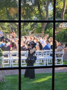 Snohomish Wedding Photographer GSquared Weddings A wedding photographer stands on grass taking photos of guests seated on white chairs outdoors, seen through a window with a wooden frame. Sunlight filters through trees in the background. Serving Seattle, Snohomish and Orlando