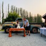 Snohomish Wedding Photographer GSquared Weddings A man and woman stand by a table cutting a cake outdoors, with an old blue pickup truck and tall trees in the background. The table is covered with an orange cloth and decorated with flowers. Serving Seattle, Snohomish and Orlando