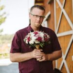 Snohomish Wedding Photographer GSquared Weddings A man wearing glasses and a maroon shirt stands outdoors by a wooden barn, holding a bouquet of pink and white flowers and looking at the camera. Serving Seattle, Snohomish and Orlando