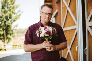 Snohomish Wedding Photographer GSquared Weddings A man wearing glasses and a maroon shirt stands outdoors by a wooden barn, holding a bouquet of pink and white flowers and looking at the camera. Serving Seattle, Snohomish and Orlando