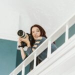 Snohomish Wedding Photographer GSquared Weddings A woman with long dark hair holds a professional camera and points it downward while standing on an indoor balcony, making a playful facial expression. She wears a blue shirt with cutout sleeves. Serving Seattle, Snohomish and Orlando