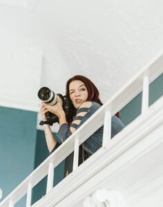 Snohomish Wedding Photographer GSquared Weddings A woman with long dark hair holds a professional camera and points it downward while standing on an indoor balcony, making a playful facial expression. She wears a blue shirt with cutout sleeves. Serving Seattle, Snohomish and Orlando