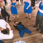 Snohomish Wedding Photographer GSquared Weddings A group of people dressed in blue formal attire dance energetically on a wooden floor while a photographer, also in blue, lies on the ground taking photos. Serving Seattle, Snohomish and Orlando