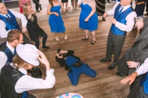 Snohomish Wedding Photographer GSquared Weddings A group of people dressed in blue formal attire dance energetically on a wooden floor while a photographer, also in blue, lies on the ground taking photos. Serving Seattle, Snohomish and Orlando