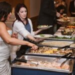 Snohomish Wedding Photographer GSquared Weddings A bride in a white dress and guests serve themselves food from a buffet table at a wedding reception, choosing from various trays of hot dishes and salads. Serving Seattle, Snohomish and Orlando