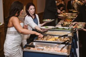 Snohomish Wedding Photographer GSquared Weddings A bride in a white dress and guests serve themselves food from a buffet table at a wedding reception, choosing from various trays of hot dishes and salads. Serving Seattle, Snohomish and Orlando