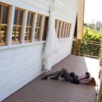 Snohomish Wedding Photographer GSquared Weddings A woman lies on a wooden deck floor, photographing a white wedding dress hanging on the exterior wall of a building with multiple windows. Sunlight brightens the scene, and greenery is visible in the background. Serving Seattle, Snohomish and Orlando
