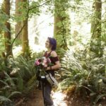 Snohomish Wedding Photographer GSquared Weddings A smiling person with purple hair stands in a sunlit forest holding a bouquet of flowers. They wear a medical walking boot, sneakers, grey pants, and a burgundy top, surrounded by lush green ferns and tall trees. Serving Seattle, Snohomish and Orlando