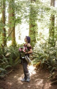 Snohomish Wedding Photographer GSquared Weddings A smiling person with purple hair stands in a sunlit forest holding a bouquet of flowers. They wear a medical walking boot, sneakers, grey pants, and a burgundy top, surrounded by lush green ferns and tall trees. Serving Seattle, Snohomish and Orlando