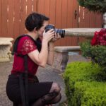 Snohomish Wedding Photographer GSquared Weddings A person wearing a red blouse, black skirt, and red heels kneels on one knee outdoors, taking a photo with a camera aimed at red flowers beside a green bush and stone benches. Serving Seattle, Snohomish and Orlando