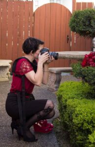 Snohomish Wedding Photographer GSquared Weddings A person wearing a red blouse, black skirt, and red heels kneels on one knee outdoors, taking a photo with a camera aimed at red flowers beside a green bush and stone benches. Serving Seattle, Snohomish and Orlando