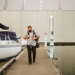Snohomish Wedding Photographer GSquared Weddings A man with a backpack walks along a dock next to a sailboat, with calm water and a large metal building in the background. Serving Seattle, Snohomish and Orlando