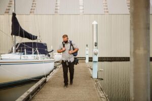 Snohomish Wedding Photographer GSquared Weddings A man with a backpack walks along a dock next to a sailboat, with calm water and a large metal building in the background. Serving Seattle, Snohomish and Orlando