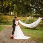 Snohomish Wedding Photographer GSquared Weddings A bride and groom kiss on a gravel path in a green park as a person holds the bride’s veil out behind her, creating a flowing effect. A bouquet of flowers lies on the ground near the couple. Serving Seattle, Snohomish and Orlando