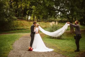 Snohomish Wedding Photographer GSquared Weddings A bride and groom kiss on a gravel path in a green park as a person holds the bride’s veil out behind her, creating a flowing effect. A bouquet of flowers lies on the ground near the couple. Serving Seattle, Snohomish and Orlando